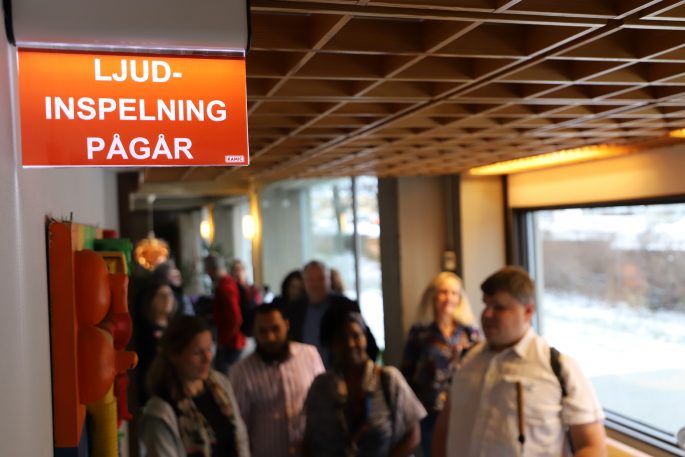 I förgrunden ljusskylten med texten Ljudinspelning pågår. Vi ser suddiga ansikten på deltagarna vid invigningen av ljudstudion i bakgrunden