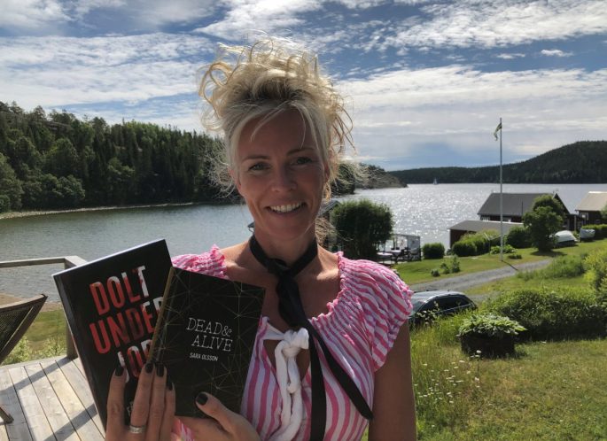 Sara har blond uppsatt hår och en rosa blus. Hon håller upp en av sina böcker. I bakgrunden ser vi Bönhamn och viken