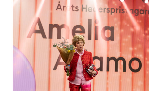 Amedlia på scenn med blommor och pris klädd organgefärgad dress och rosa blus